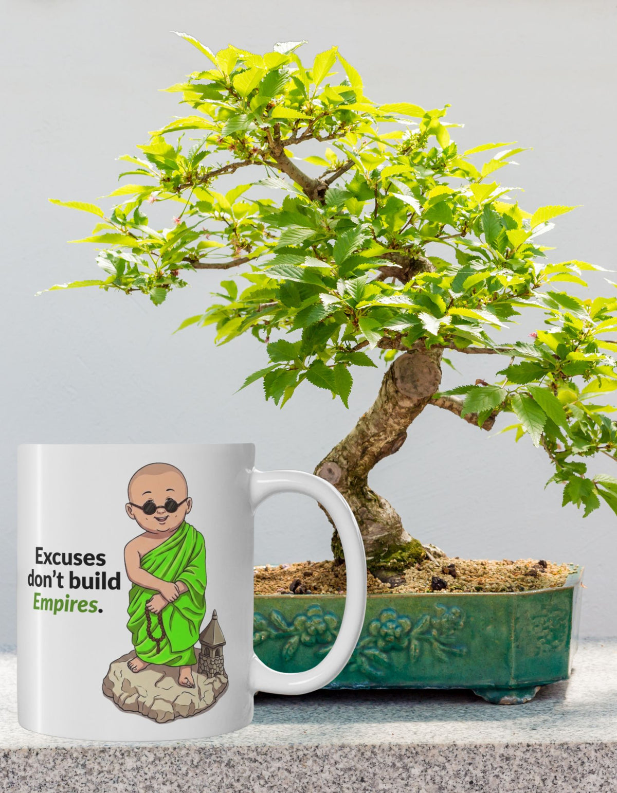 A white ceramic mug with an illustration of a monk wearing green robes and the text 'Excuses don't build Empires' is shown next to a potted bonsai tree on a grey stone surface against a light grey background.