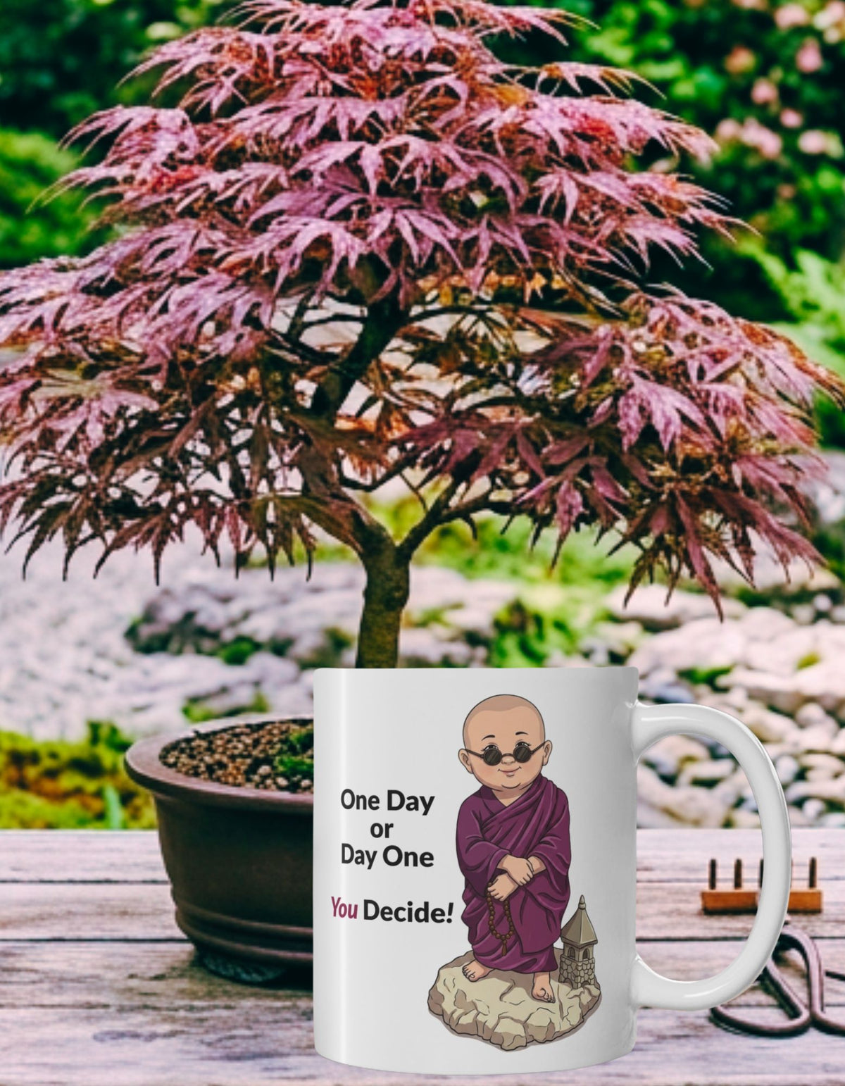 A white ceramic mug with an illustration of a monk in a purple robe, accompanied by the text 'One Day or Day One You Decide!'. The mug is placed on a wooden table in front of a potted red-leafed Japanese maple tree, with a blurred garden background.