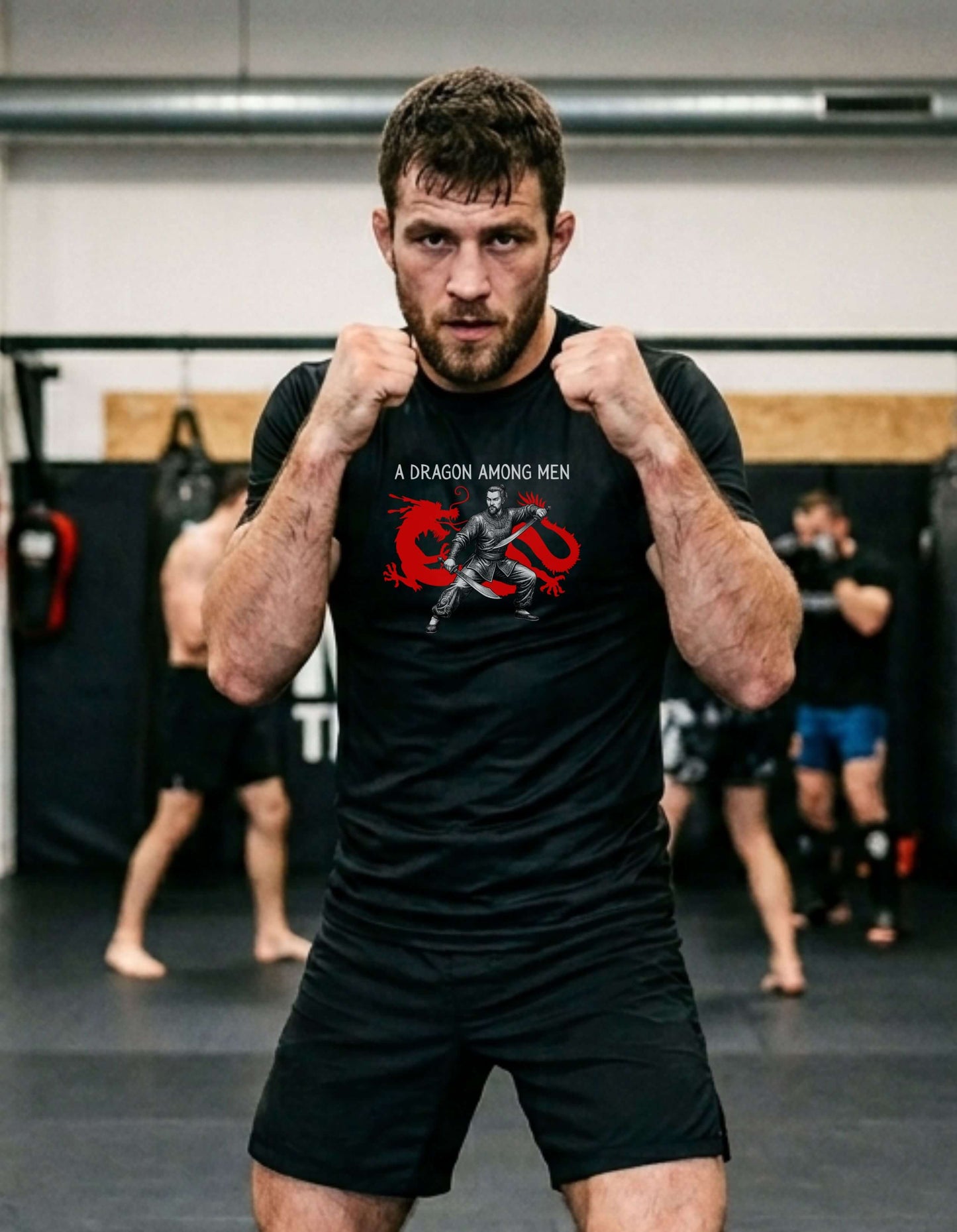 Man in a MMA gym fists up, wearing a black t-shirt with the slogan - A Dragon among Men printed in white, above the bold graphic of a male warrior in fighting stance wielding dual swords in front of a red dragon silhouette.