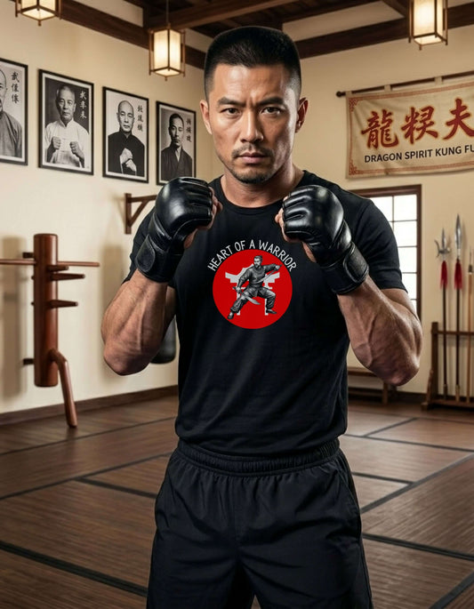 Man in a martial arts gym fists up, wearing a black t-shirt with the slogan - Heart of a Warrior printed in white, above a red circle featuring the bold graphic of a male warrior in fighting stance wielding dual swords in front of a white Chinese temple.