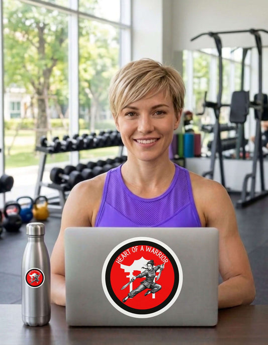 Fit woman in a gym setting with a laptop  and water bottle each featuring a 'Heart of a Warrior' sticker.