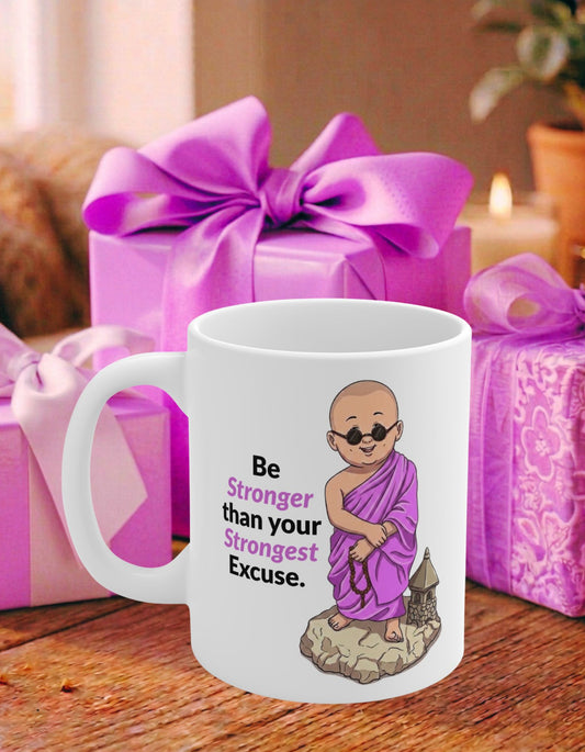 A white ceramic mug with an illustration of a monk in pink-purple robes and the text 'Be Stronger than your Strongest Excuse.' It is placed on a wooden table in front of pink gift boxes with pink bows.