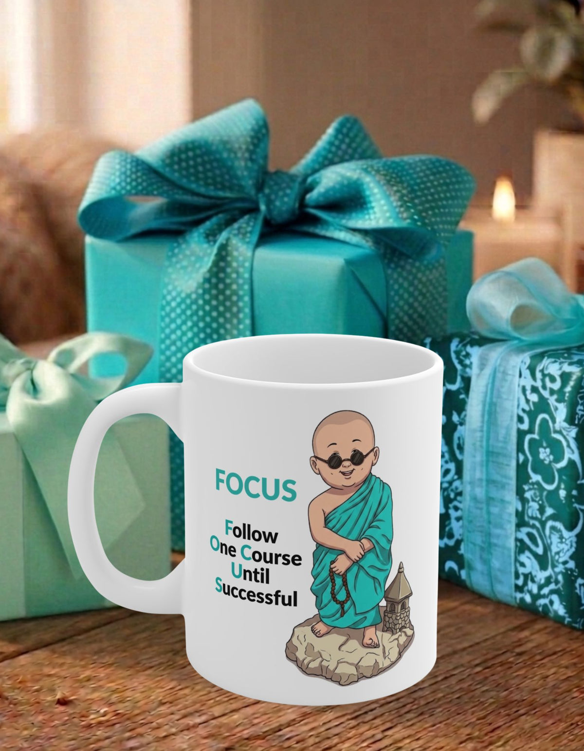 A white ceramic coffee mug with an illustration of a monk and the text 'FOCUS - Follow One Course Until Successful' in teal and black. The mug is set on a wooden table with teal gift boxes in the background.
