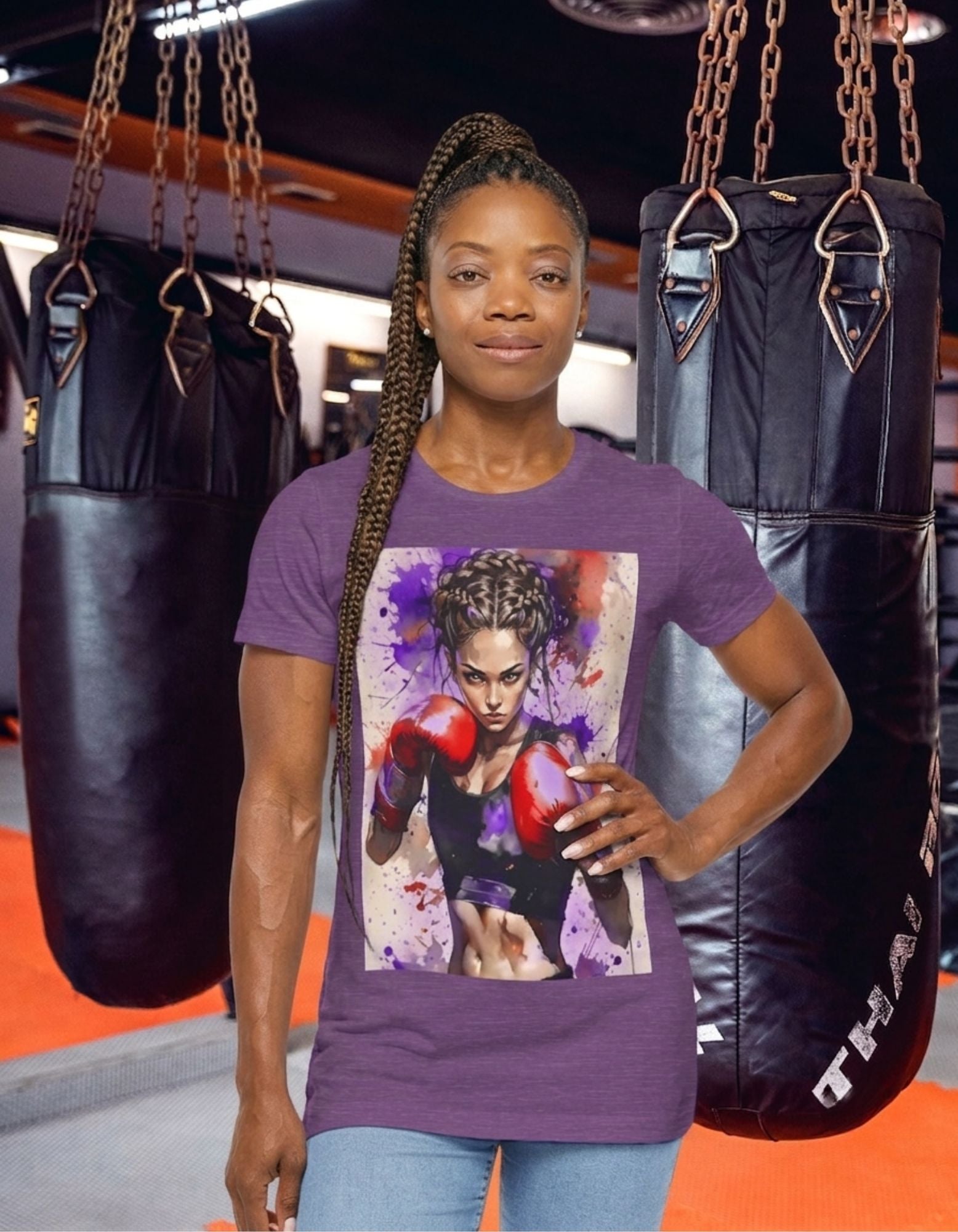 A woman stands in a gym wearing a heather team purple t-shirt with a boxer girl design, flanked by two black hanging punching bags.