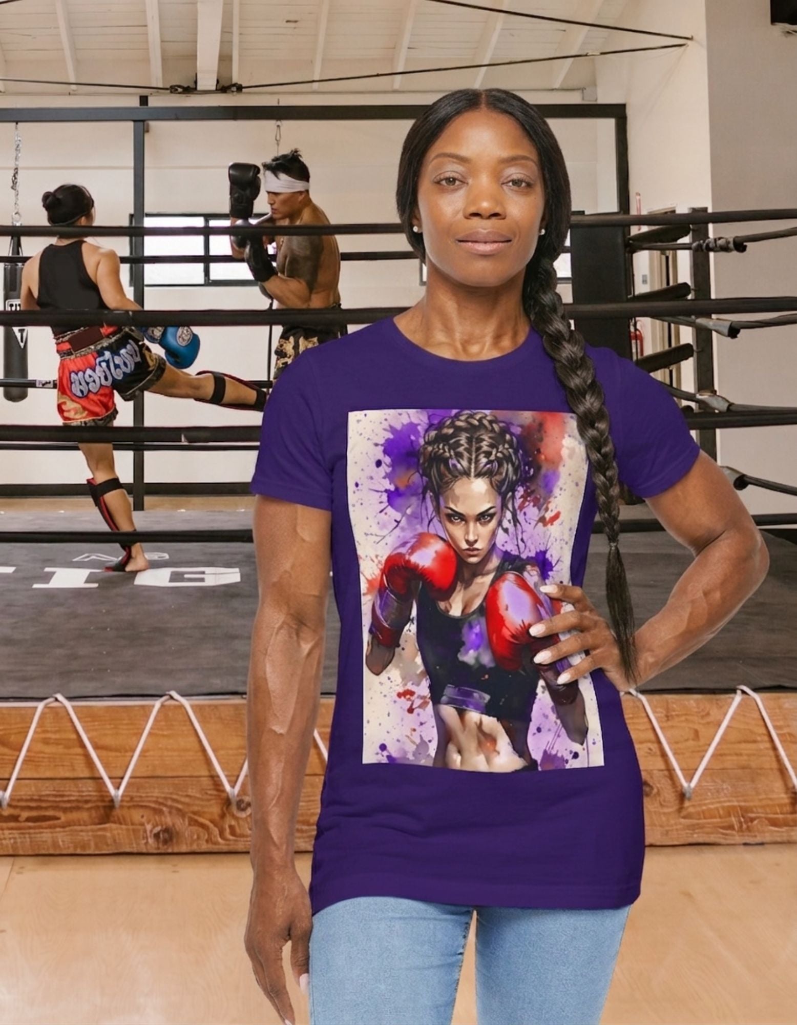 A woman wearing a team purple t-shirt with a boxer girl design, stands in front of a boxing ring with two fighters fighting in it.