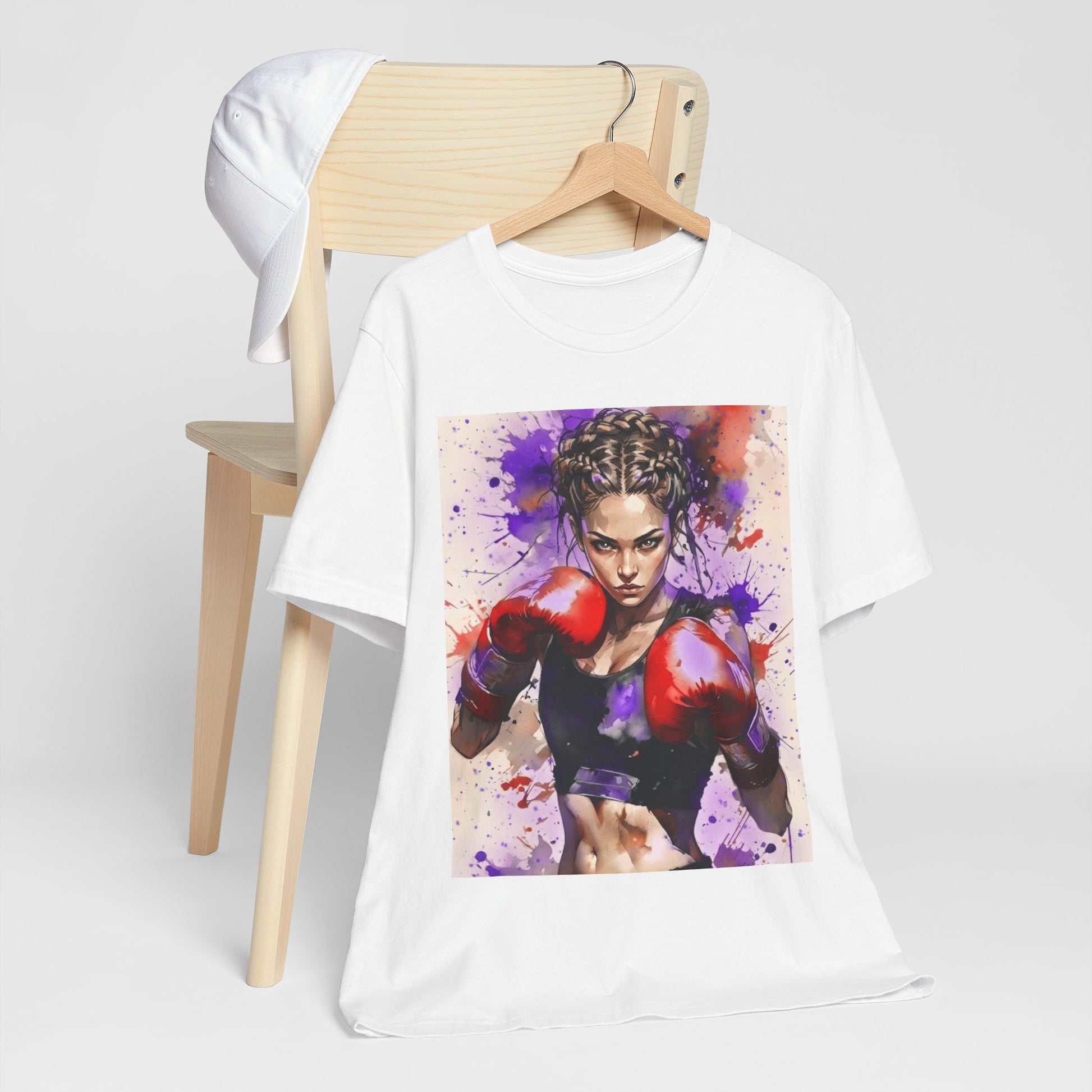 A white short-sleeve t-shirt on a wooden hanger, displayed in front of a light wood chair. The t-shirt features a large rectangular graphic print of a female boxer wearing red boxing gloves against a purple and pink paint splatter background.