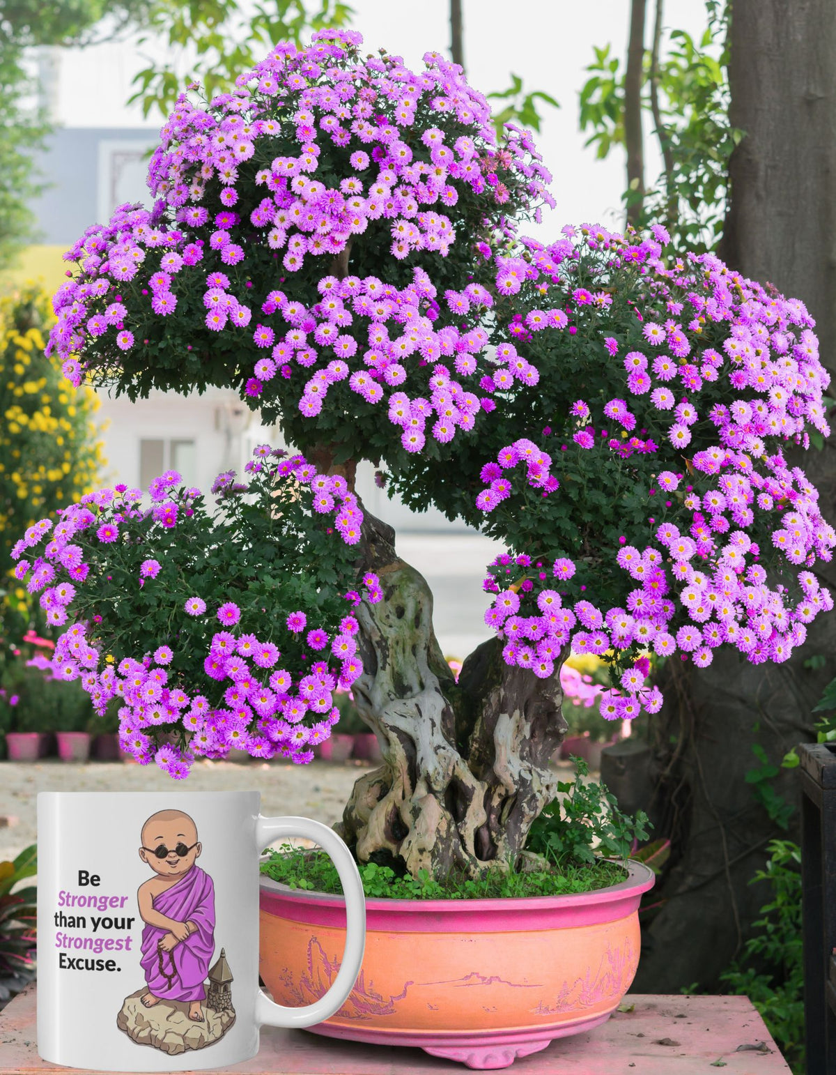 A white ceramic mug with an  illustration of a monk wearing pink-purple robes and the text 'Be stronger than your strongest excuse.' Behind the mug is a blooming bonsai-style plant with dense pink-purple flowers, shown outdoors in a peach-coloured pot.