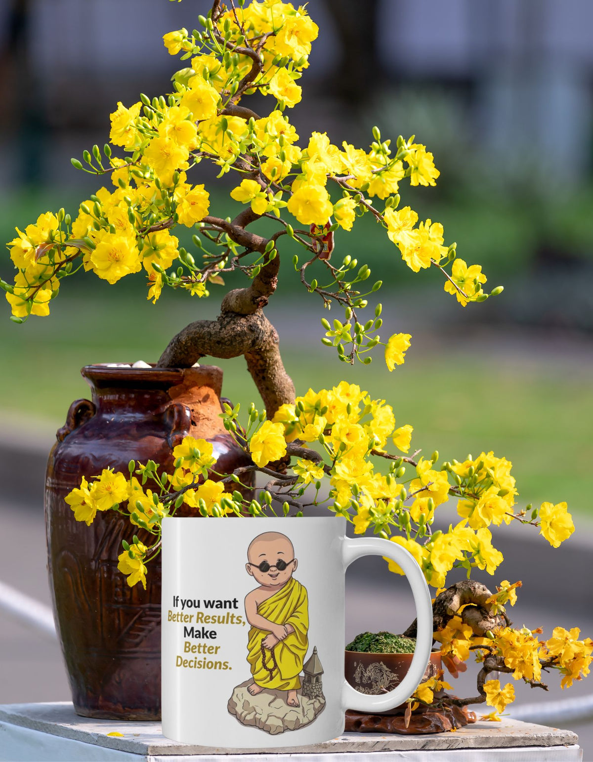 A white ceramic mug with an illustration of a monk in yellow robes and the text 'If you want Better Results Make Better Decisions.' placed in front of a potted bonsai tree with bright yellow flowers.