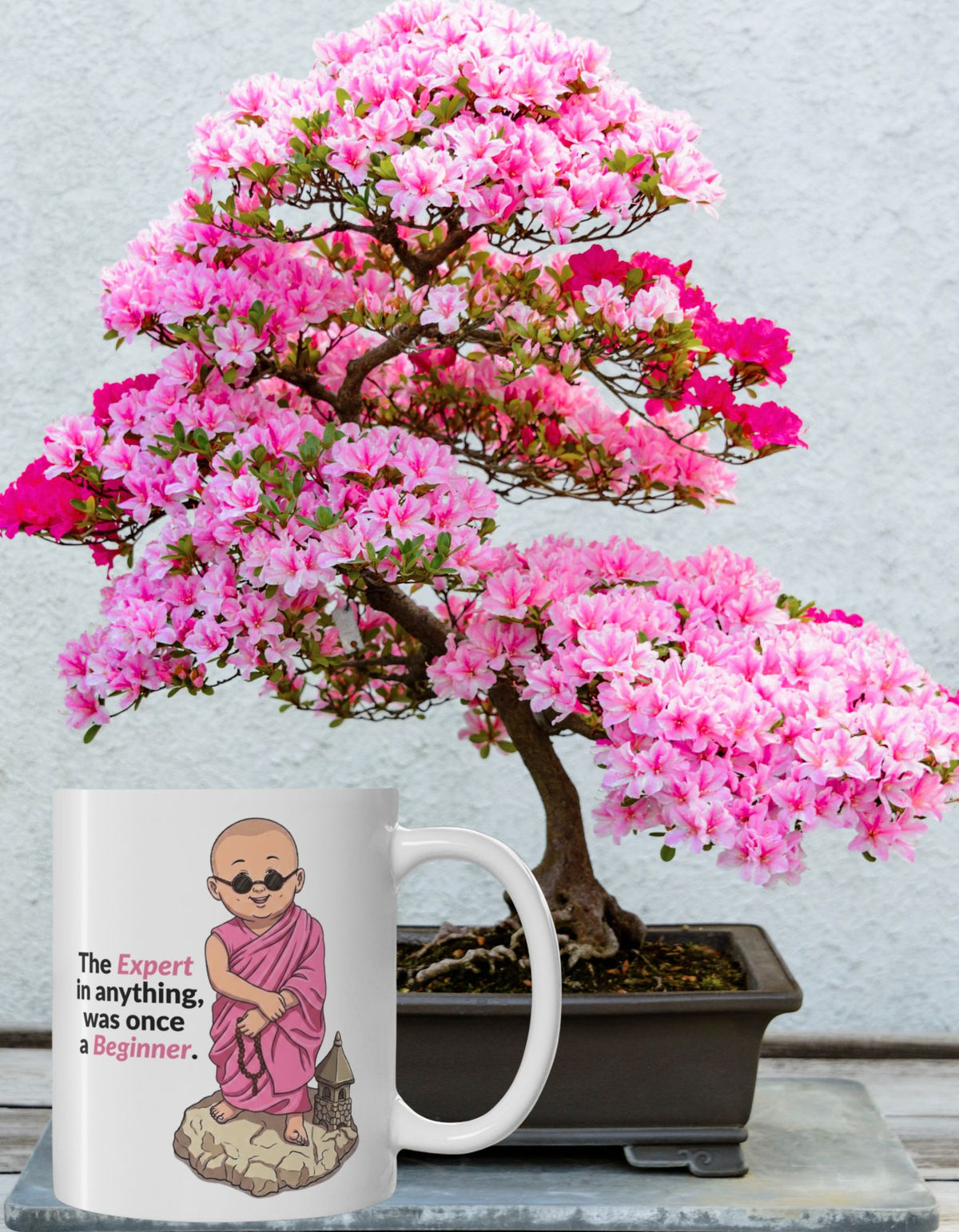 A white ceramic mug with an illustration of a monk wearing pink robes next to the text 'The expert in anything was once a beginner.' on a stone slab with a pink flower bonsai tree in the background.
