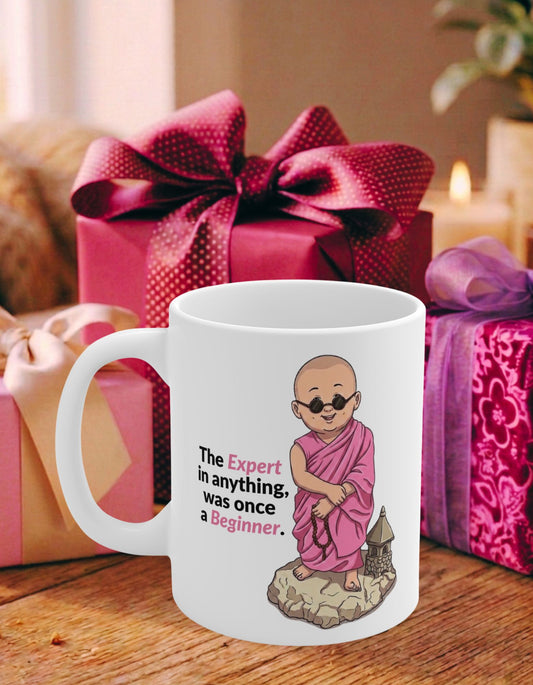 A white ceramic mug with an illustration of a monk in pink robes next to the text 'The expert in anything was once a beginner.' on a table with pink wrapped gifts in the background.