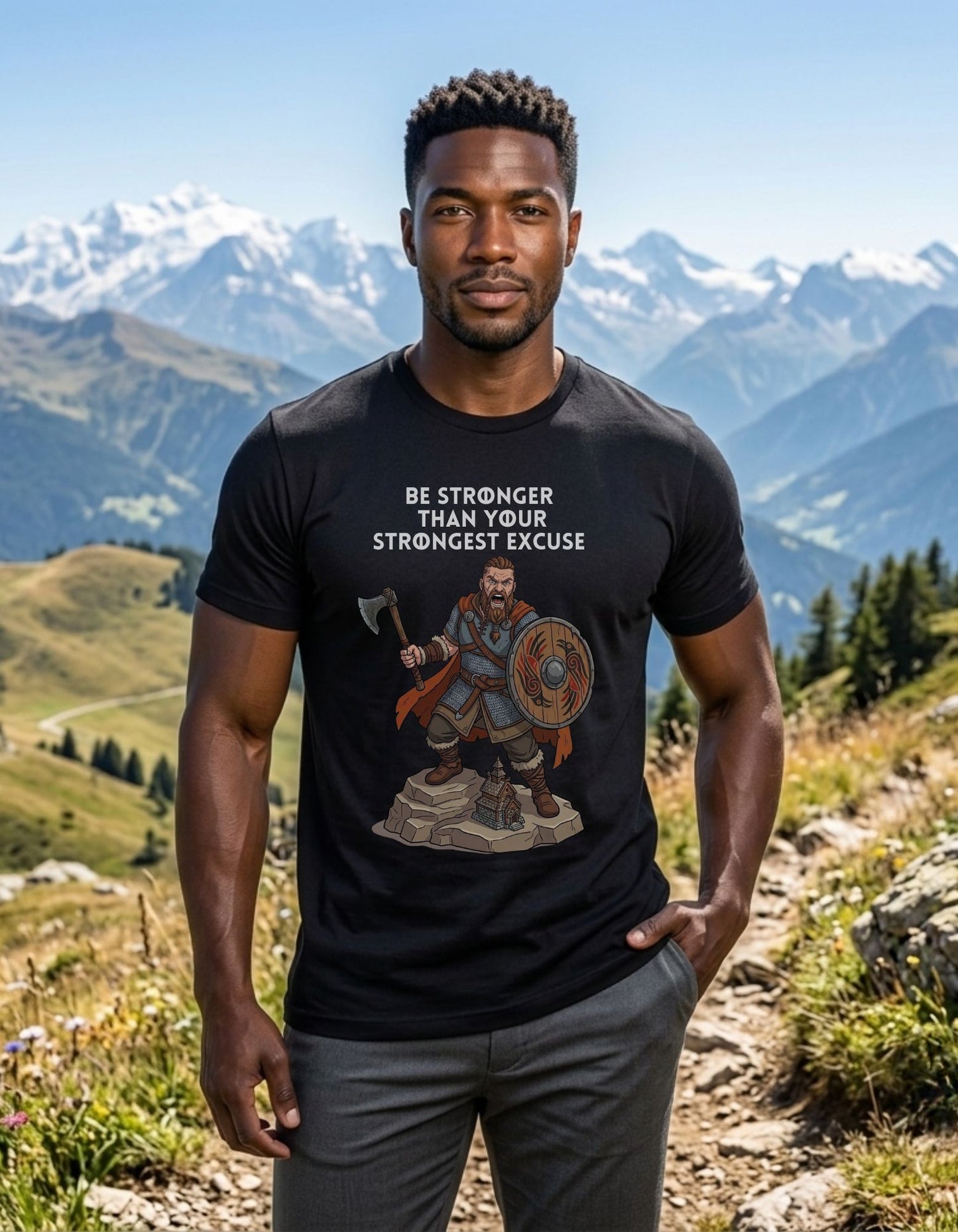 A man standing outdoors in a mountain landscape wearing a black graphic t-shirt with a Viking warrior holding an axe and shield illustration and the text 'BE STRONGER THAN YOUR STRONGEST EXCUSE'.