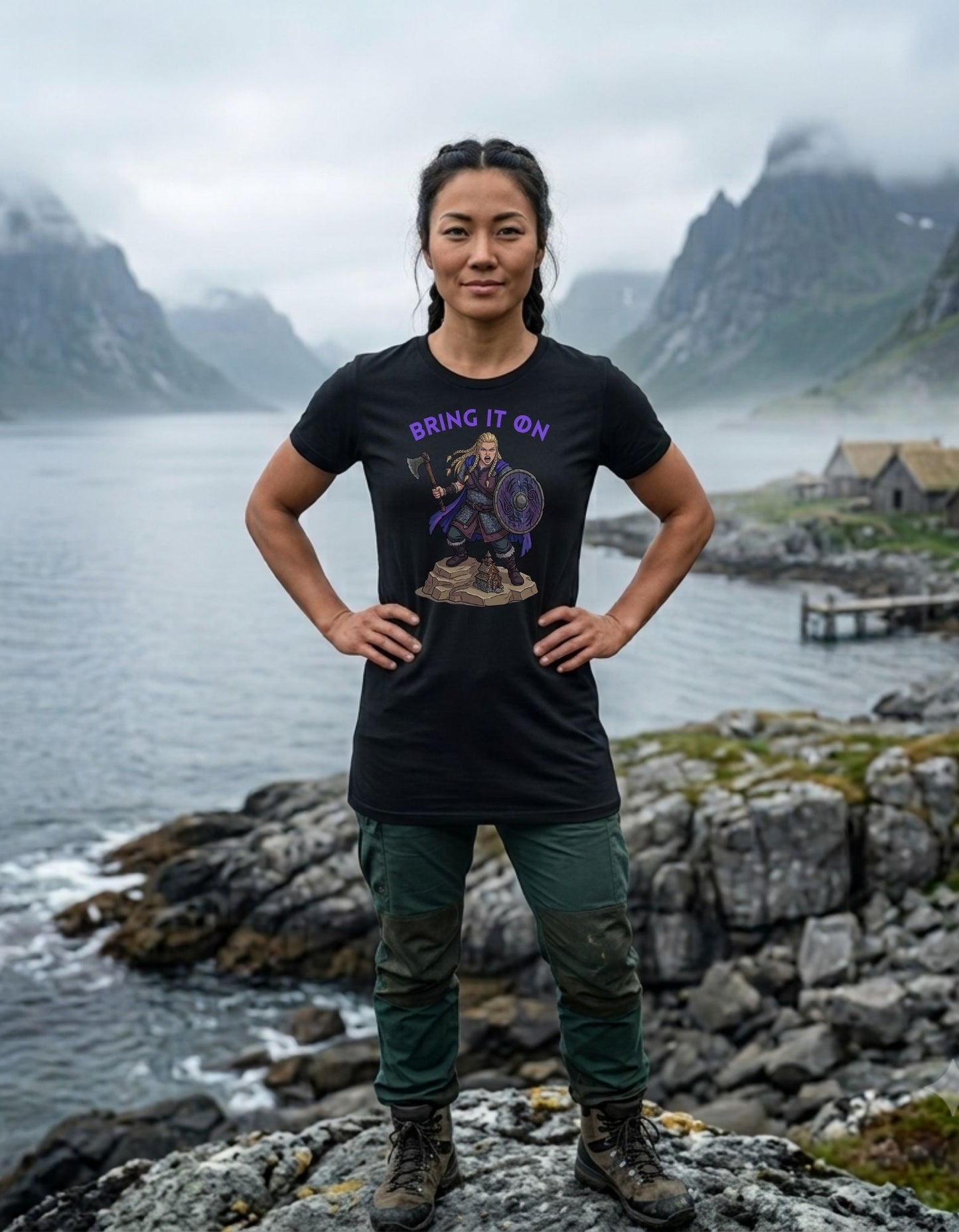 A woman stands outdoors on a rocky shoreline with water, mountains, and a small building in the background, wearing a black graphic T-shirt with purple text reading 'BRING IT ON' with a colourful Viking woman warrior design beneath the text.