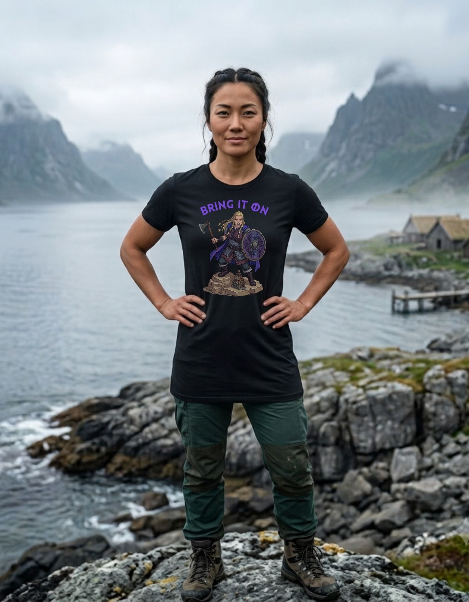 A woman stands outdoors on a rocky shoreline with water, mountains, and a small building in the background, wearing a black graphic T-shirt with purple text reading 'BRING IT ON' with a colourful Viking woman warrior design beneath the text.
