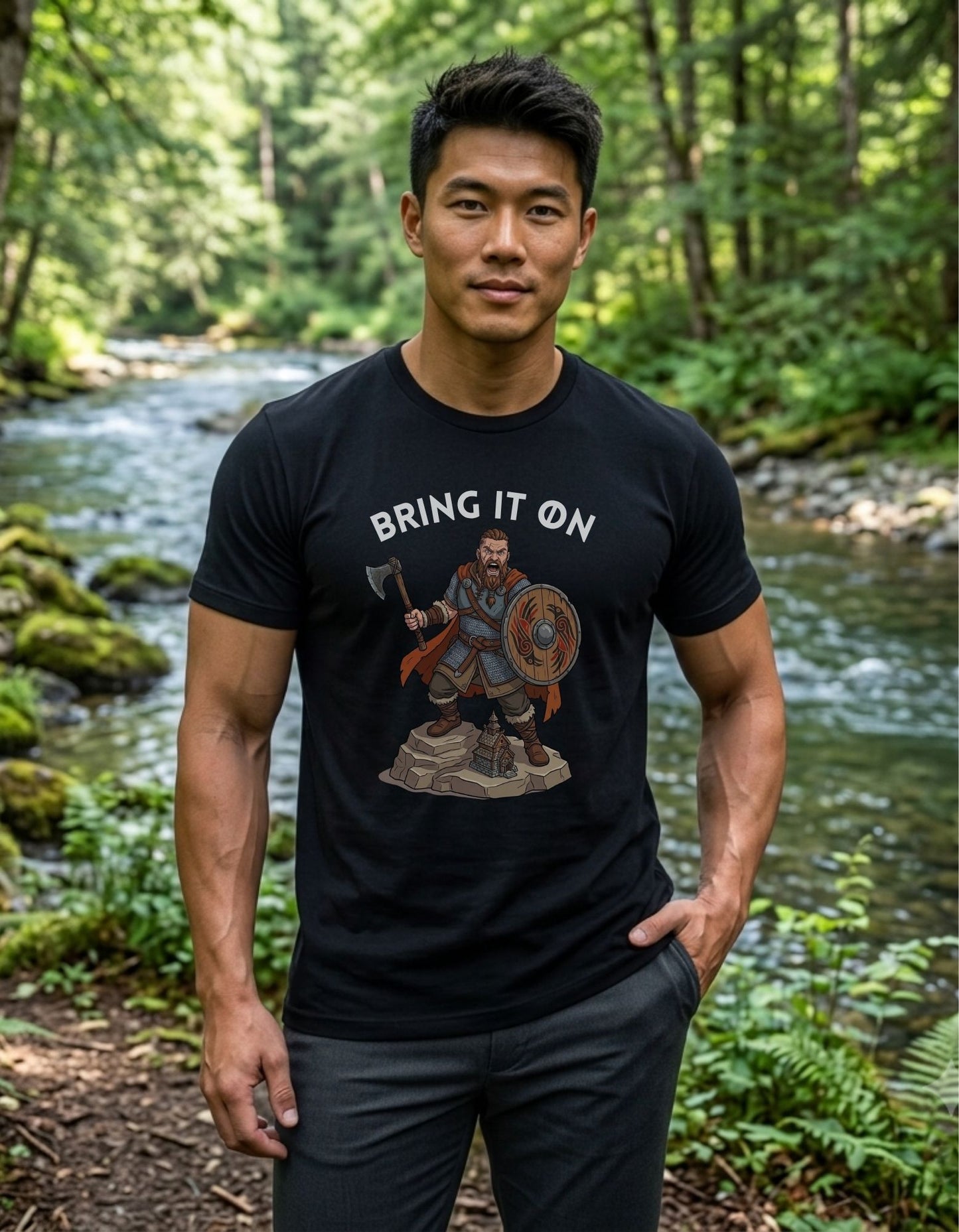 A man standing outdoors in a forest by a flowing stream, wearing a black graphic T-shirt with the text 'BRING IT ON' above an illustration of a Viking warrior holding an axe and shield.