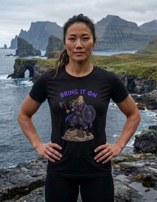 A woman stands outdoors on a rocky coastal cliff with water and dramatic mountains in the background, wearing a black graphic T-shirt. The shirt has purple text reading 'BRING IT ON' above a printed illustration of a female Viking warrior holding an axe and shield.