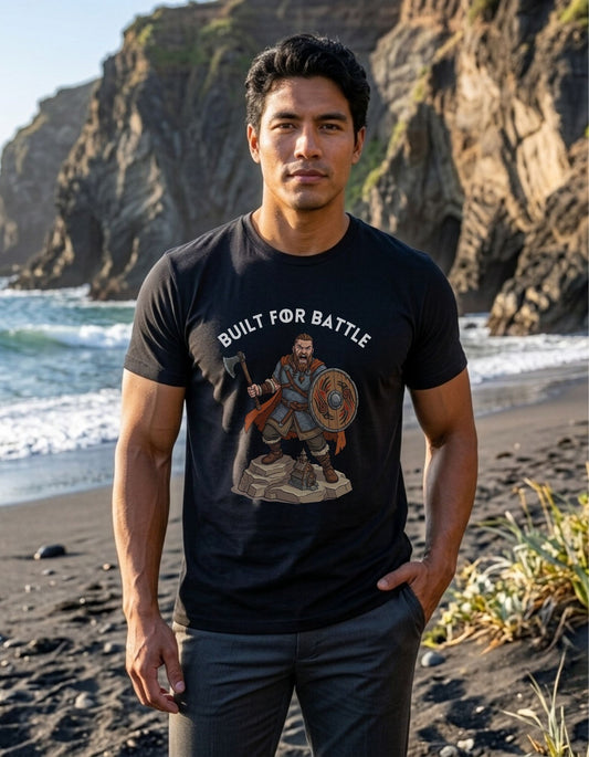 A man standing on a beach wearing a black graphic t-shirt with a Viking warrior holding an axe and shield illustration below the text 'BUILT FOR BATTLE'.