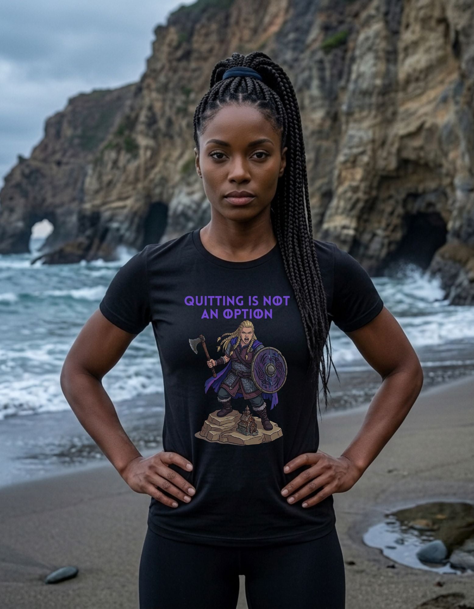 A woman stands on a beach with rocky cliffs and ocean waves behind her, wearing a black graphic T-shirt with the text 'QUITTING IS NOT AN OPTION' and a printed illustration of a female Viking warrior holding a shield and axe.