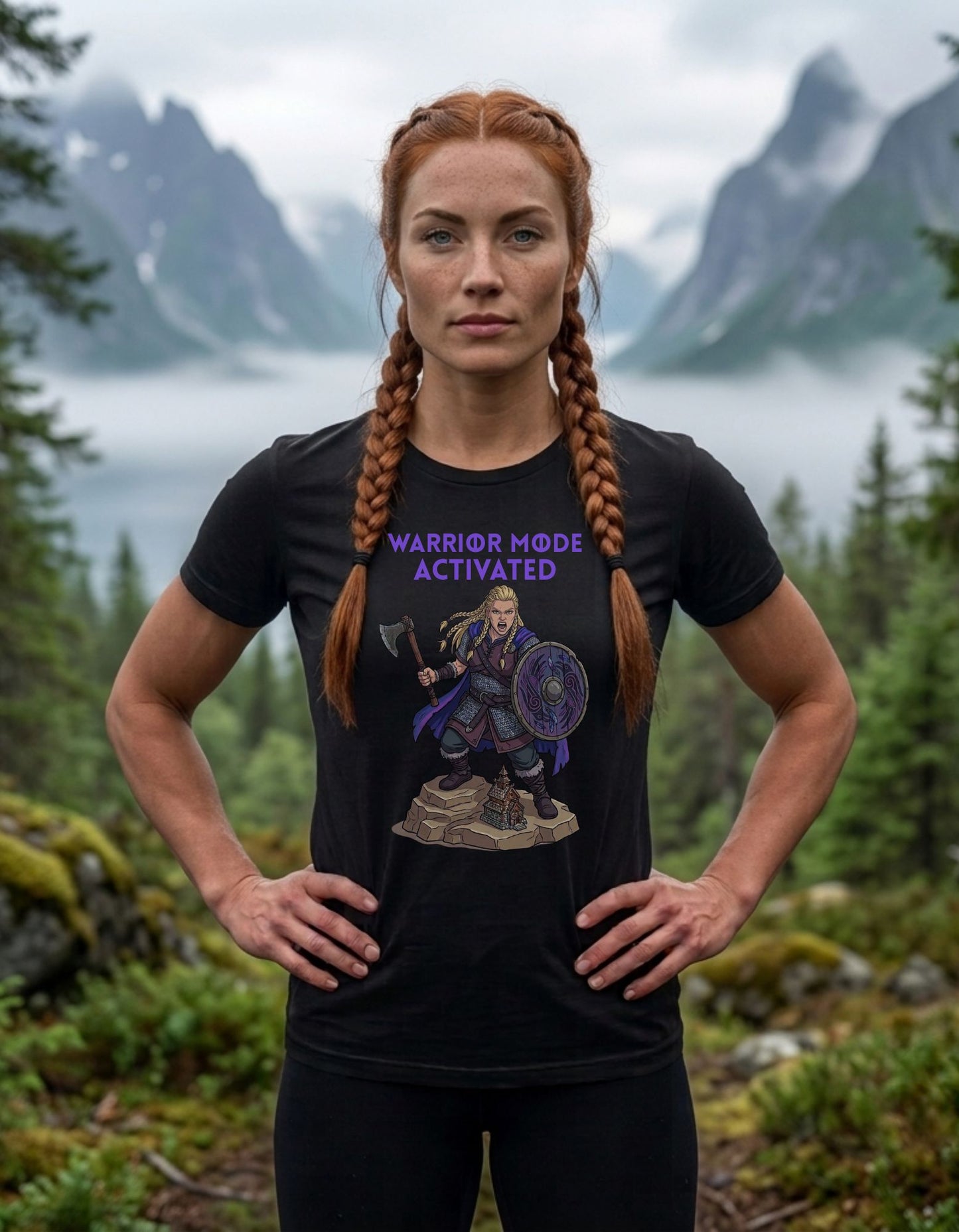 A woman stands outdoors in a mountain landscape wearing a black graphic T-shirt with purple text reading 'WARRIOR MODE ACTIVATED' and a female Viking warrior illustration.