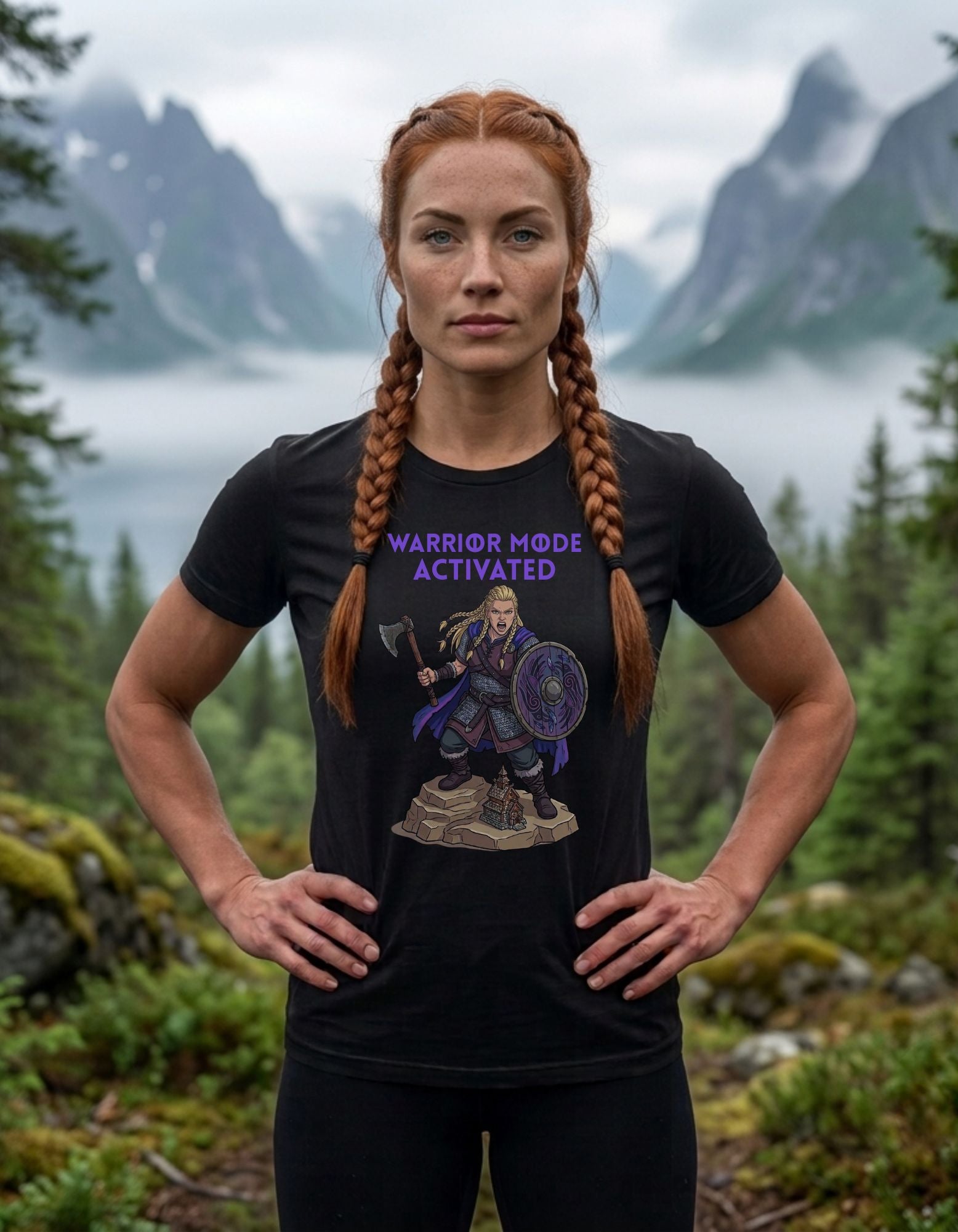 A woman stands outdoors in a mountain landscape wearing a black graphic T-shirt with purple text reading 'WARRIOR MODE ACTIVATED' and a female Viking warrior illustration.