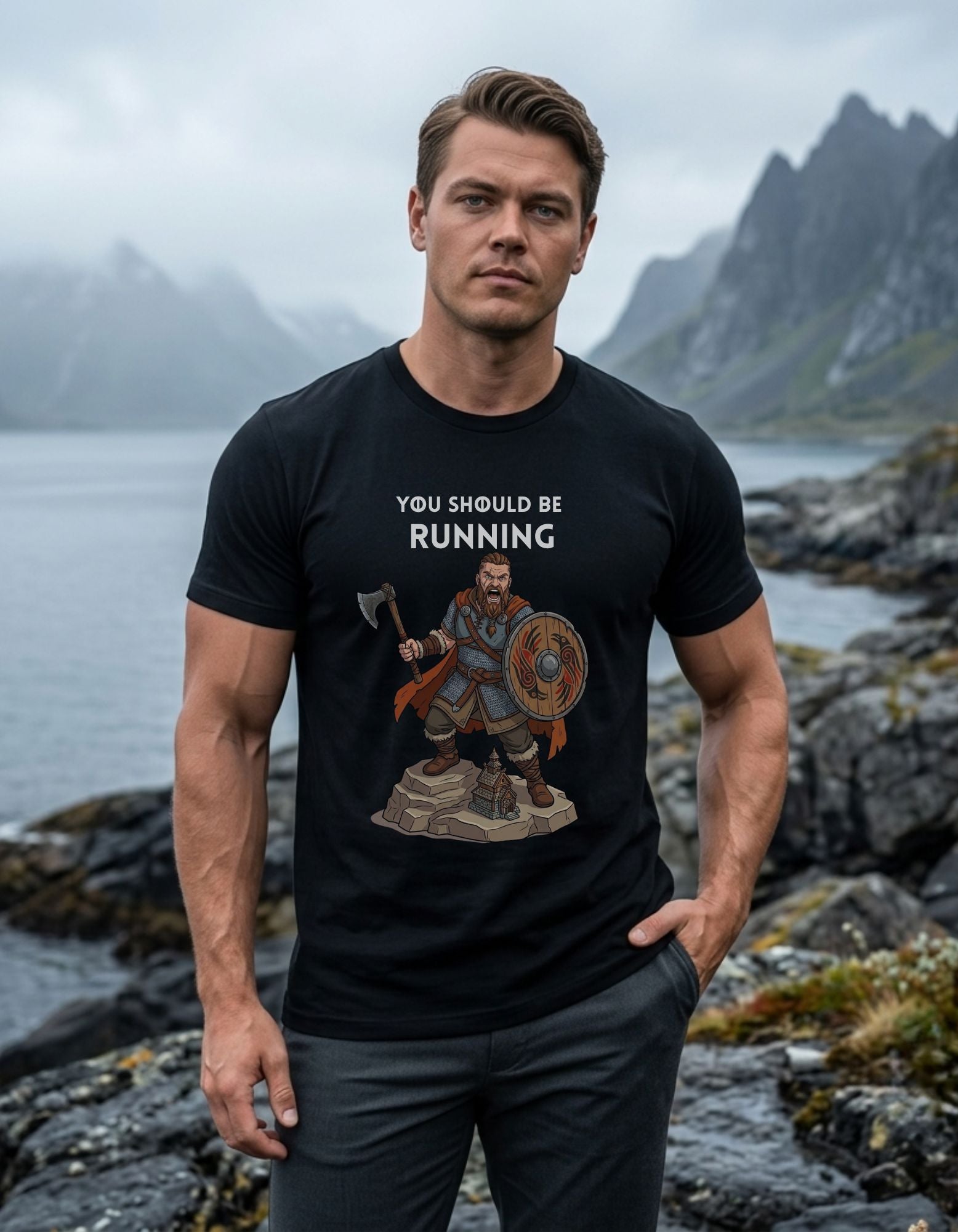 A muscular man wearing a black graphic t-shirt stands on a rocky shoreline with misty mountains and water in the background. The t-shirt features a white text graphic reading 'YOU SHOULD BE RUNNING' above an illustration of a Viking warrior holding an axe and wearing a shield.